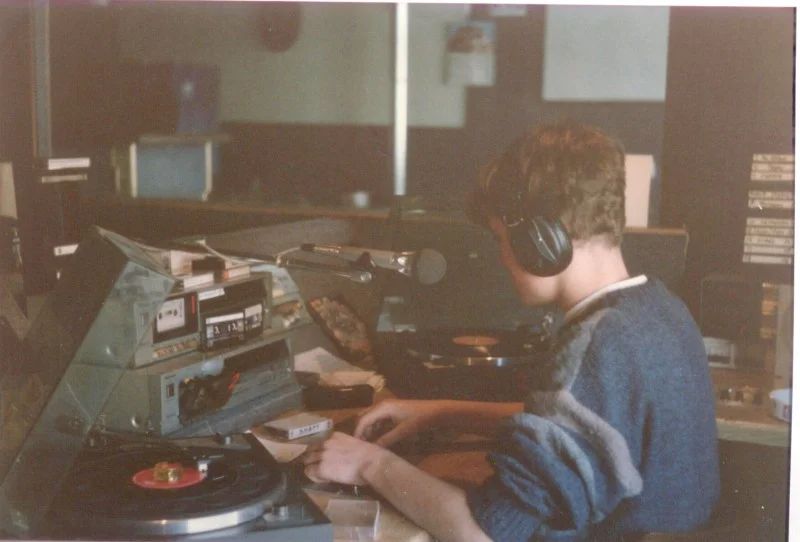 Dj met koptelefoon van achteren gezien achter draaitafels, cassettedeck en microfoon in een vrije radiostudio, jaren tachtig